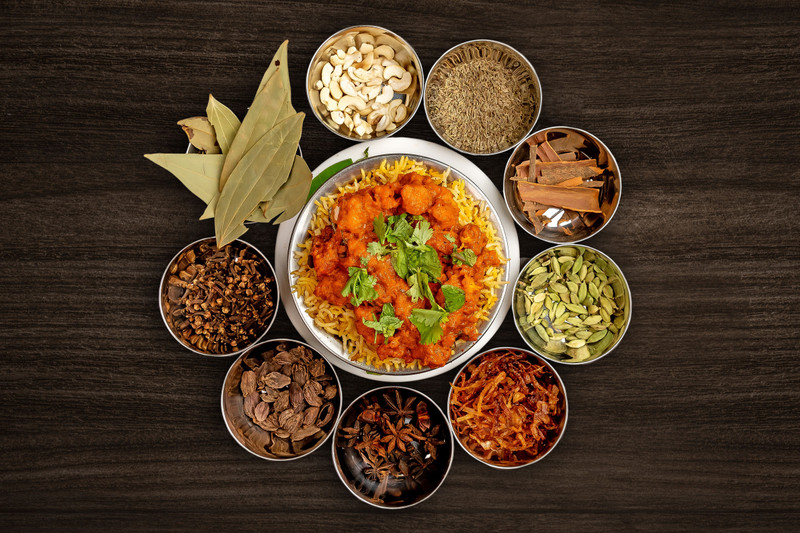 Bowls of spices around a curry on dark wood.